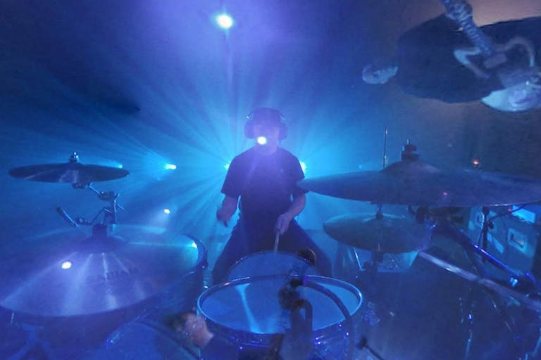Fish-eye lens of a man sat at a drum kit with other band members circled around, light up blue with spotlights behind them