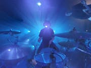 Fish-eye lens of a man sat at a drum kit with other band members circled around, light up blue with spotlights behind them