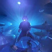 Fish-eye lens of a man sat at a drum kit with other band members circled around, light up blue with spotlights behind them