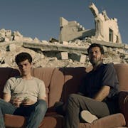 A Palestinian teen boy and older man sit on torn sofa amongst the rubble of a bombed home