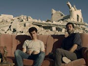 A Palestinian teen boy and older man sit on torn sofa amongst the rubble of a bombed home