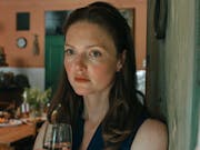 A white woman with red hair and freckles stands in a middle class kitchen holding a glass of wine looking haunted
