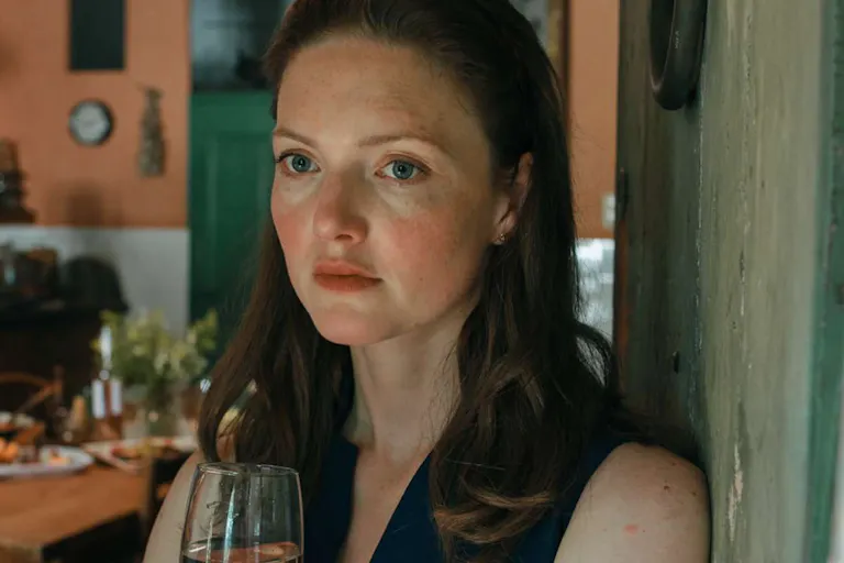 A white woman with red hair and freckles stands in a middle class kitchen holding a glass of wine looking haunted