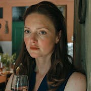 A white woman with red hair and freckles stands in a middle class kitchen holding a glass of wine looking haunted
