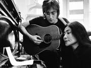 Black and white still of a white man with small round glasses playing the guitar next to a Japanese woman sitting playing a piano