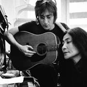 Black and white still of a white man with small round glasses playing the guitar next to a Japanese woman sitting playing a piano