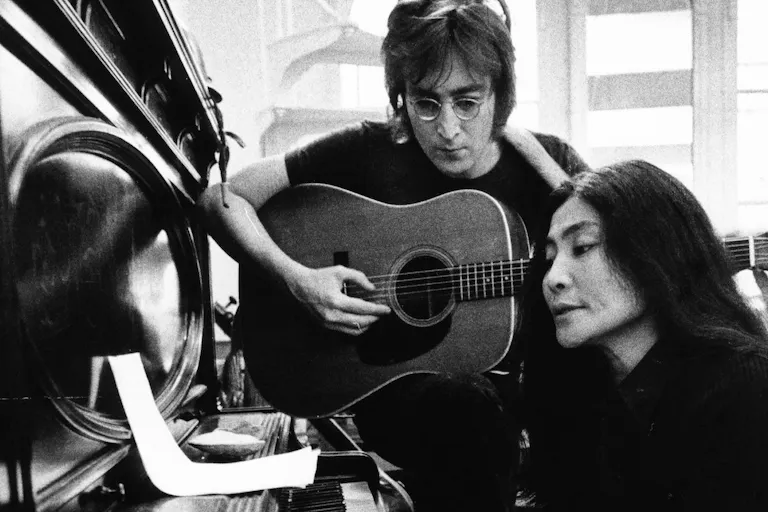 Black and white still of a white man with small round glasses playing the guitar next to a Japanese woman sitting playing a piano