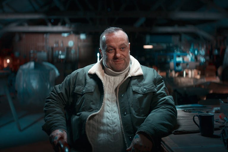 A white middle aged man wearing a jumper and jacket, sitting at a dark workshop table with a smirk on his face