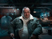 A white middle aged man wearing a jumper and jacket, sitting at a dark workshop table with a smirk on his face