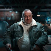 A white middle aged man wearing a jumper and jacket, sitting at a dark workshop table with a smirk on his face