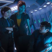 A white woman, a blue alien woman, a Black man, a brown man and a white man, crowd around a spaceship console desk