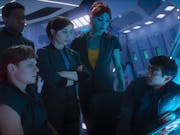 A white woman, a blue alien woman, a Black man, a brown man and a white man, crowd around a spaceship console desk