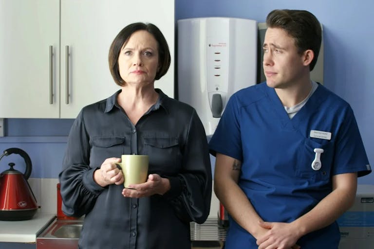 A white woman and a young white man in blue scrubs stand in a hospital break room