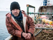 A white middle aged man in a thick waterproof coat, jumper and beanie hat, on a small fishing vessel