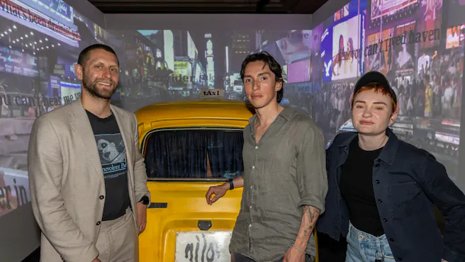 two men and one woman stand in front of a model taxi in a room with screens on each wall