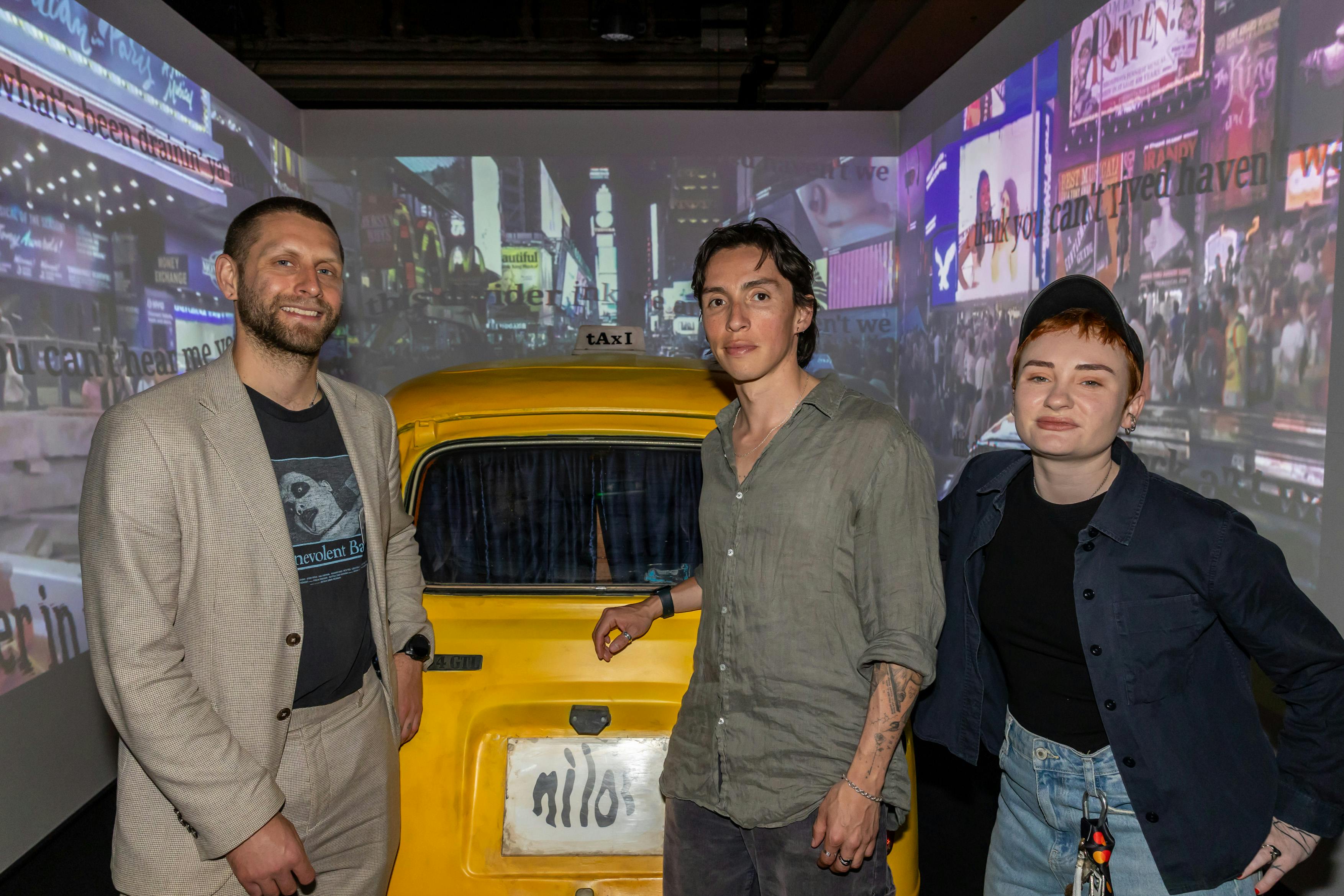 two men and one woman stand in front of a model taxi in a room with screens on each wall