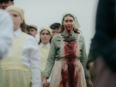 A young white woman in a pinafore with blood on her face and down her dress, surrounded by other woman in similar clothes