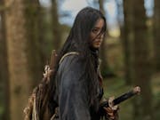 A young Japanese woman with long dark hair blowing across her face stands in a forest with a sword at her hip