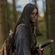 A young Japanese woman with long dark hair blowing across her face stands in a forest with a sword at her hip