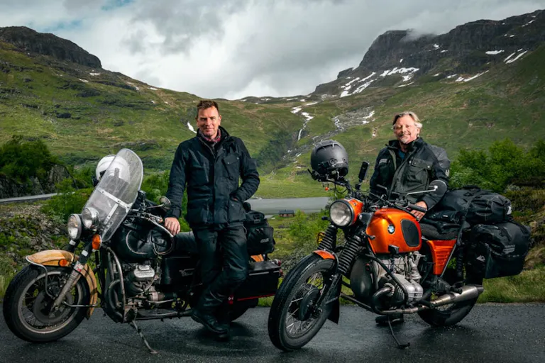 Two middle aged white men beside their motorcycles with a picturesque green mountain behind them