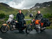 Two middle aged white men beside their motorcycles with a picturesque green mountain behind them