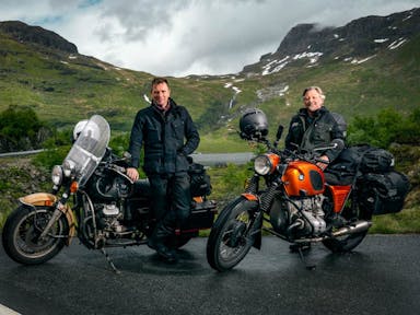 Two middle aged white men beside their motorcycles with a picturesque green mountain behind them