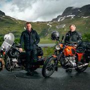 Two middle aged white men beside their motorcycles with a picturesque green mountain behind them