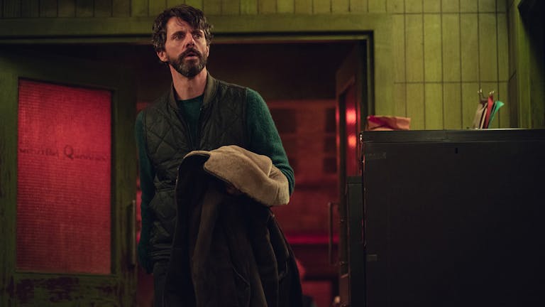 A middle aged white man with dark hair and beard in a dark room looking concerned