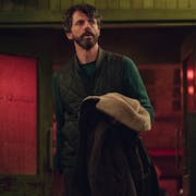 A middle aged white man with dark hair and beard in a dark room looking concerned