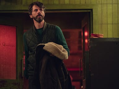 A middle aged white man with dark hair and beard in a dark room looking concerned