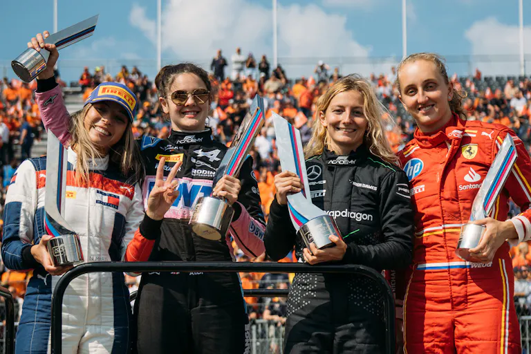 Four white women Formula 1 drivers holding up trophies as a crowd cheers behind them