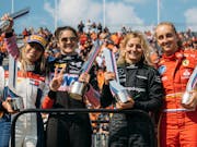 Four white women Formula 1 drivers holding up trophies as a crowd cheers behind them