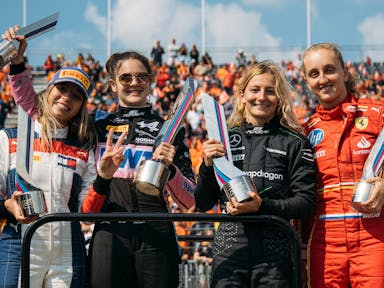 Four white women Formula 1 drivers holding up trophies as a crowd cheers behind them