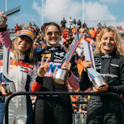 Four white women Formula 1 drivers holding up trophies as a crowd cheers behind them