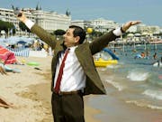 A white man in a tweed jacket and red tie holding a digital camera aloft on a beach in Cannes