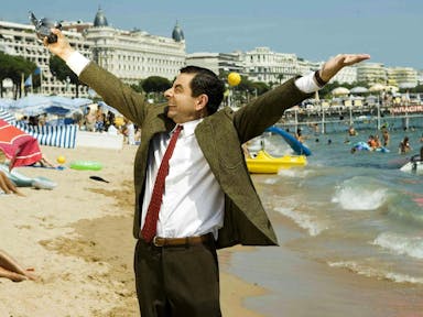 A white man in a tweed jacket and red tie holding a digital camera aloft on a beach in Cannes