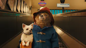 Live action with a CGI brown bear in a blue duffle coat and red hat holding a white dog coming down the escalators in Paddington station