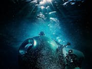 A small commercial submarine in the dark, gloomy ocean with a strip of blue light illuminating it from above