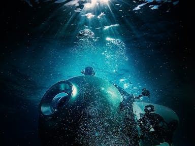 A small commercial submarine in the dark, gloomy ocean with a strip of blue light illuminating it from above