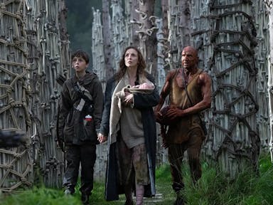 A white woman holding a baby next to a young white boy and an older white man covered in blood standing in front of pillars of bones