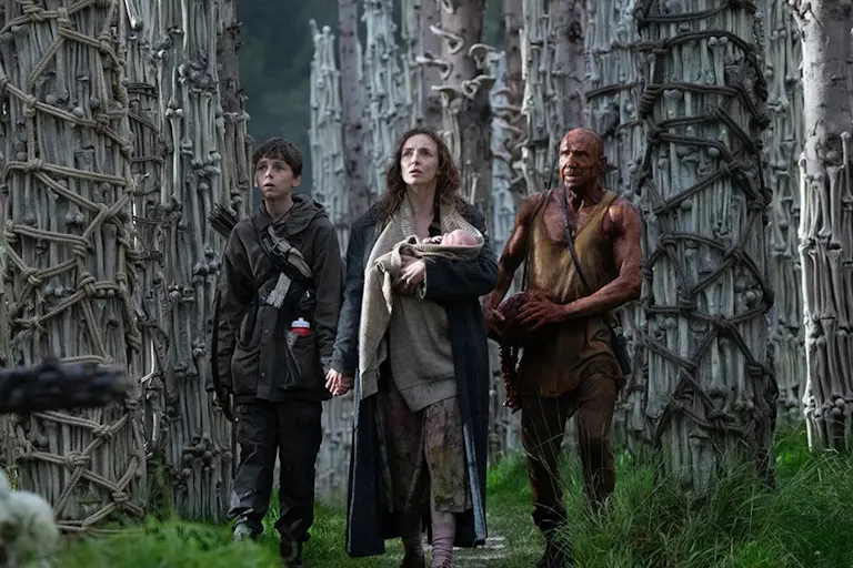 A white woman holding a baby next to a young white boy and an older white man covered in blood standing in front of pillars of bones
