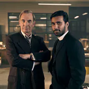 A middle aged white man in a casual suit and a younger brown man in vicar's clothes standing in a 60s police office