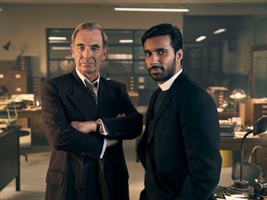 A middle aged white man in a casual suit and a younger brown man in vicar's clothes standing in a 60s police office