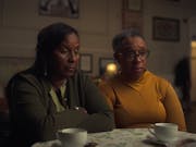 A middle aged Black woman and an older Black woman sitting in a low lit room looking haunted