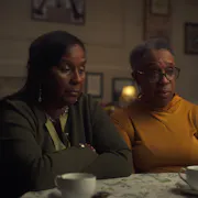A middle aged Black woman and an older Black woman sitting in a low lit room looking haunted