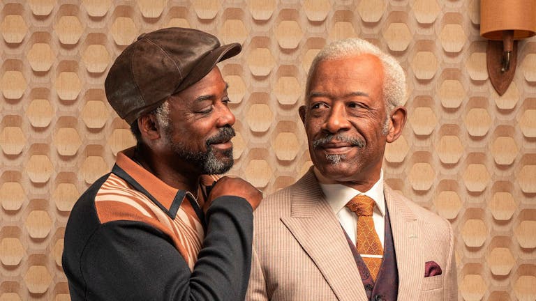 Two older Black men with grey hair and beards, one wearing a light coloured suit, the other in a hat and jumper with his arm around the other