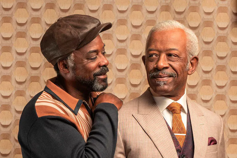 Two older Black men with grey hair and beards, one wearing a light coloured suit, the other in a hat and jumper with his arm around the other