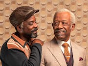 Two older Black men with grey hair and beards, one wearing a light coloured suit, the other in a hat and jumper with his arm around the other