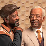Two older Black men with grey hair and beards, one wearing a light coloured suit, the other in a hat and jumper with his arm around the other