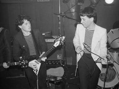 Black and white still of two young white men on a small stage singing and playing the guitar in the 80s